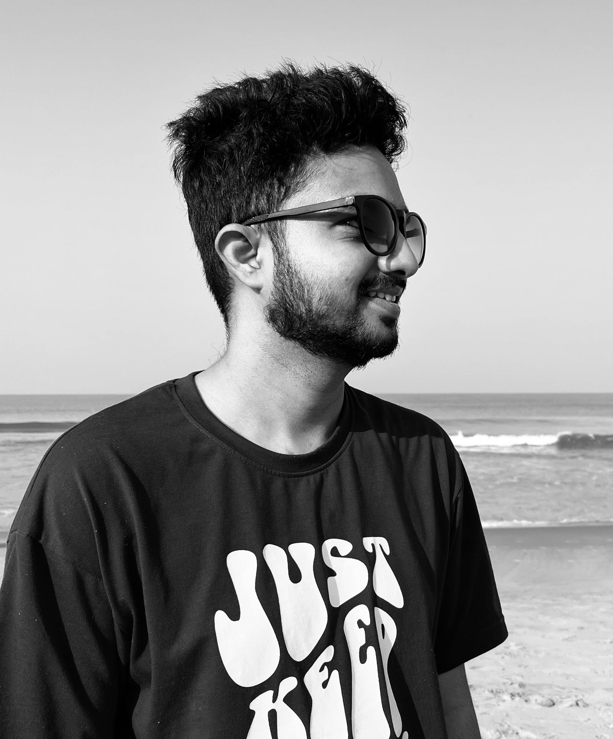 Black and white photo of a young man wearing sunglasses at the beach with calm waves in the background.